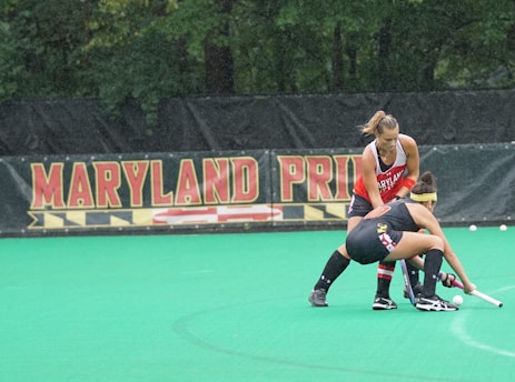 Two female field hockey players, one wearing a red jersey and the other wearing a black jersey with yellow and red accents, are engaged in a play on a green field. They are both focused on controlling the ball, with one player bending down with her stick while the other stands close behind. A banner in the background displays the words 'Maryland Pride'. Trees form the backdrop beyond the banner.