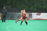 A female athlete in a red and black uniform plays field hockey on a green field. Rain is visible during the game, and spectators or coaches wearing jackets are present on the sidelines.