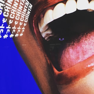 A close-up image of an open mouth with visible teeth and a prominent tongue. The lips are colored with a deep red shade, contrasting with the white teeth. The background features a blue color with text, including the word 'CHINTAI' repeated multiple times in white, along with other symbols.
