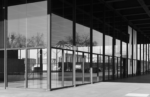 A modern architectural structure with large glass panels reflecting a cityscape, trees, and part of a classical building. The design features clean lines and a grid-like pattern, emphasizing the blend of contemporary and traditional elements.