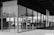 A modern architectural structure with large glass panels reflecting a cityscape, trees, and part of a classical building. The design features clean lines and a grid-like pattern, emphasizing the blend of contemporary and traditional elements.