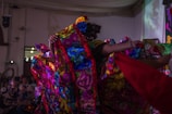 A vibrant scene depicting a traditional dance performance with colorful, floral-patterned costumes. The performers are energetically moving, creating dynamic and flowing patterns with their outfits in a warmly lit indoor setting.