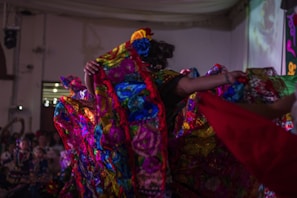 A vibrant scene depicting a traditional dance performance with colorful, floral-patterned costumes. The performers are energetically moving, creating dynamic and flowing patterns with their outfits in a warmly lit indoor setting.
