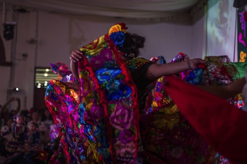 A vibrant scene depicting a traditional dance performance with colorful, floral-patterned costumes. The performers are energetically moving, creating dynamic and flowing patterns with their outfits in a warmly lit indoor setting.