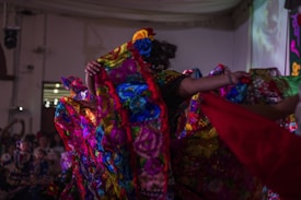 A vibrant scene depicting a traditional dance performance with colorful, floral-patterned costumes. The performers are energetically moving, creating dynamic and flowing patterns with their outfits in a warmly lit indoor setting.