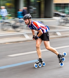 A person rollerblading at high speed on a paved road, wearing a helmet and a sporty outfit. The background is slightly blurred, suggesting swift motion, and includes a railing and some bicycles.