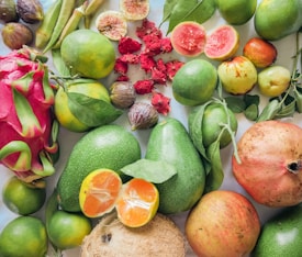 A vibrant assortment of fresh fruits and vegetables, including pomegranates, dragon fruit, figs, guavas, and avocados, arranged on a light surface. The produce is varied, with a mix of green, red, orange, and pink hues, showcasing their natural textures and colors.