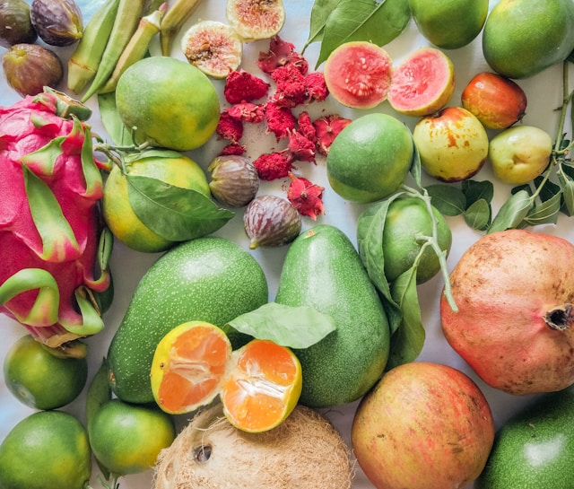 A vibrant assortment of fresh fruits and vegetables, including pomegranates, dragon fruit, figs, guavas, and avocados, arranged on a light surface. The produce is varied, with a mix of green, red, orange, and pink hues, showcasing their natural textures and colors.