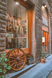 A rustic storefront featuring hanging cured meats inside the window, a large wooden wagon wheel leaning against the glass, and a small chalkboard sign near some potted plants. The exterior is crafted with stonework, and there's an inviting entrance to a restaurant.