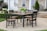 A wooden patio dining set with black chairs and a matching table sits on a concrete patio. A pitcher of lemonade and a potted plant are on the table. The background features a house with stone and siding exterior, white columns, and a neatly trimmed garden with hydrangeas and a potted evergreen.