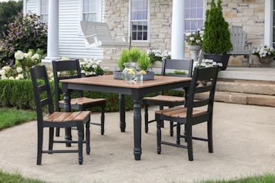 A wooden patio dining set with black chairs and a matching table sits on a concrete patio. A pitcher of lemonade and a potted plant are on the table. The background features a house with stone and siding exterior, white columns, and a neatly trimmed garden with hydrangeas and a potted evergreen.