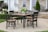 A wooden patio dining set with black chairs and a matching table sits on a concrete patio. A pitcher of lemonade and a potted plant are on the table. The background features a house with stone and siding exterior, white columns, and a neatly trimmed garden with hydrangeas and a potted evergreen.