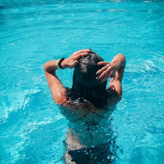 A person with long dark hair stands waist-deep in a swimming pool, holding their hair with both hands. The water is clear and blue, reflecting sunlight.