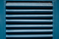 A close-up of a blue louvered vent with horizontal slats showing the texture of painted wood. The paint appears slightly worn with visible signs of weathering. There are small traces of dust or spider webs in the corners.