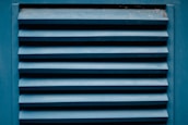 A close-up of a blue louvered vent with horizontal slats showing the texture of painted wood. The paint appears slightly worn with visible signs of weathering. There are small traces of dust or spider webs in the corners.