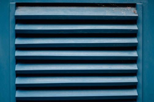 A close-up of a blue louvered vent with horizontal slats showing the texture of painted wood. The paint appears slightly worn with visible signs of weathering. There are small traces of dust or spider webs in the corners.