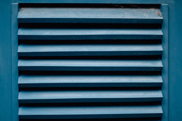 A close-up of a blue louvered vent with horizontal slats showing the texture of painted wood. The paint appears slightly worn with visible signs of weathering. There are small traces of dust or spider webs in the corners.