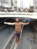 A man is seen performing slackline walking over an urban road with a crowd of onlookers gathered on a bridge. He is balancing confidently and appears focused. Behind him, a cyclist can be seen on the road, and three people are standing further back, observing. Tall buildings and a mixture of shaded and sunlit areas create a contrast in the backdrop.