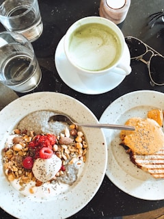 A table with a light breakfast spread consisting of a bowl of chia seed pudding topped with nuts, raspberries, and a dollop of yogurt. Next to it, a grilled sandwich with a cheese topping. Two glasses of water, a cup of green tea, and a pair of sunglasses are also present.