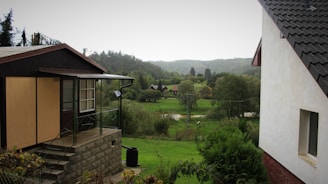 A serene rural landscape featuring two houses, one in the foreground with a stone foundation and another partially visible from the right. They are surrounded by lush green lawns, trees, and bushes. In the background, a forested area and a distant hill are visible under a cloudy sky. The scene is peaceful and appears to be in a countryside setting.