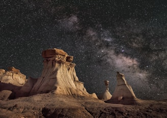 Dramatic rock formations under a vibrant, star-filled night sky with the Milky Way galaxy clearly visible in the background. The scene combines earthy desert tones with the expansive beauty of the cosmos, creating a stark contrast between the rugged terrain and the celestial panorama above.