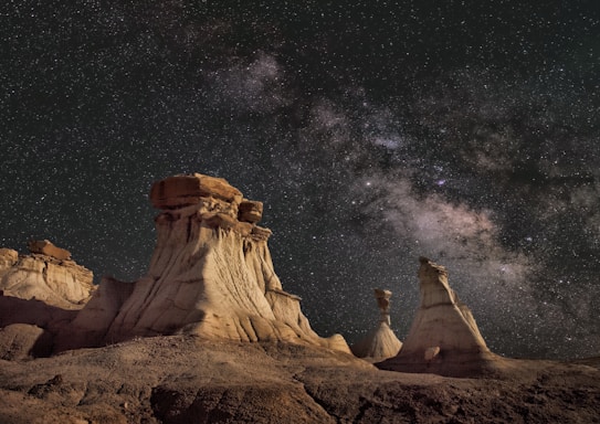 Dramatic rock formations under a vibrant, star-filled night sky with the Milky Way galaxy clearly visible in the background. The scene combines earthy desert tones with the expansive beauty of the cosmos, creating a stark contrast between the rugged terrain and the celestial panorama above.