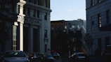 The image presents an urban street scene with several multi-story buildings. One prominent building, labeled 'First Community Bank,' is constructed in a classical style with tall columns. Shadows cast by the buildings and trees suggest the time is either early morning or late afternoon. Several cars are parked along the street, and there are bicycles visible near the sidewalk.