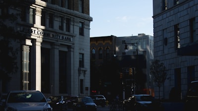 The image presents an urban street scene with several multi-story buildings. One prominent building, labeled 'First Community Bank,' is constructed in a classical style with tall columns. Shadows cast by the buildings and trees suggest the time is either early morning or late afternoon. Several cars are parked along the street, and there are bicycles visible near the sidewalk.