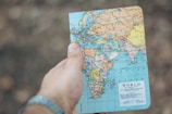 A hand holding a small world map focused on Europe, Africa, and parts of Asia. The background is blurred and earthy, suggesting an outdoor setting.