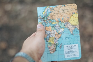 A hand holding a small world map focused on Europe, Africa, and parts of Asia. The background is blurred and earthy, suggesting an outdoor setting.
