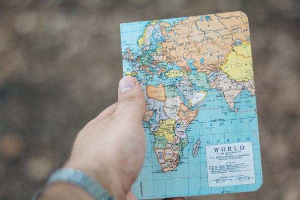 A hand holding a small world map focused on Europe, Africa, and parts of Asia. The background is blurred and earthy, suggesting an outdoor setting.