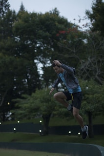 A person is captured mid-air while performing a jump in an outdoor setting. The individual wears athletic gear including a jacket, shorts, and running shoes. The background consists of lush green trees and a cloudy sky, with some lights visible near the ground.