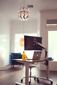 A modern, minimalist workspace with a standing desk featuring a monitor and a laptop covered in stickers. To the left is a blue armchair and a soft rug. Above the desk hangs a contemporary, spherical pendant light with geometric patterns.