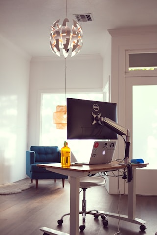 A modern, minimalist workspace with a standing desk featuring a monitor and a laptop covered in stickers. To the left is a blue armchair and a soft rug. Above the desk hangs a contemporary, spherical pendant light with geometric patterns.