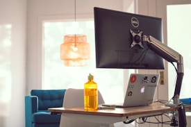 A desk setup featuring a large monitor on an adjustable arm, a laptop decorated with stickers, and a yellow water bottle. The background shows a modern room with a blue couch and a woven pendant light, with natural light streaming through a window.