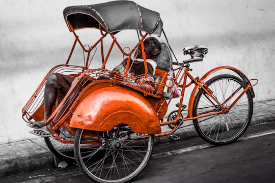 A person is resting in an orange tricycle with a black canopy, parked against a plain wall. The tricycle has a vintage design with intricate metalwork and visible wear, suggesting regular use.