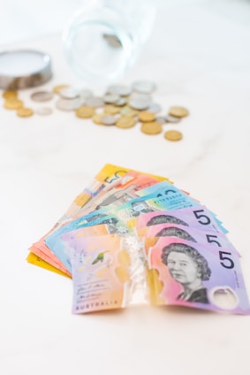 A fan of colorful Australian banknotes is spread out prominently in the foreground, featuring denominations with distinct designs. In the background, there is a transparent glass jar tipped over with various coins scattered around it on a light surface.