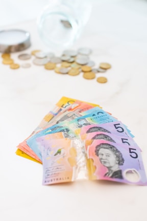 A fan of colorful Australian banknotes is spread out prominently in the foreground, featuring denominations with distinct designs. In the background, there is a transparent glass jar tipped over with various coins scattered around it on a light surface.