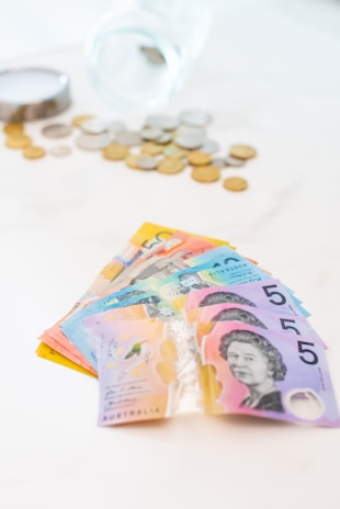 A fan of colorful Australian banknotes is spread out prominently in the foreground, featuring denominations with distinct designs. In the background, there is a transparent glass jar tipped over with various coins scattered around it on a light surface.