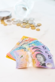 A fan of colorful Australian banknotes is spread out prominently in the foreground, featuring denominations with distinct designs. In the background, there is a transparent glass jar tipped over with various coins scattered around it on a light surface.