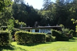 A small house is nestled in a lush green landscape surrounded by dense trees. The structure is modest, with a flat roof and a mix of small windows. The lawn in front is well-maintained, and hedges and shrubs add to the natural scenery. The forested background suggests a peaceful, secluded setting.