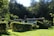 A small house is nestled in a lush green landscape surrounded by dense trees. The structure is modest, with a flat roof and a mix of small windows. The lawn in front is well-maintained, and hedges and shrubs add to the natural scenery. The forested background suggests a peaceful, secluded setting.
