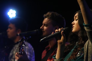 A group of three musicians performing with microphones and instruments. The lighting suggests a stage setting, focusing on the individuals as they sing or play music. The close-up perspective emphasizes the engagement and emotion of the performers.