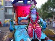 A colorful statue of a deity with blue skin, adorned with a garland of red flowers and wearing bright, ornate clothing. The background features vibrant decorations and what appears to be a peacock. The setting suggests a festive or ceremonial atmosphere.