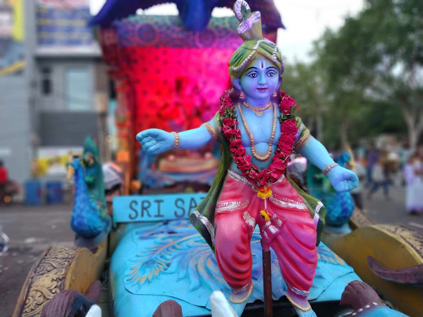 A colorful statue of a deity with blue skin, adorned with a garland of red flowers and wearing bright, ornate clothing. The background features vibrant decorations and what appears to be a peacock. The setting suggests a festive or ceremonial atmosphere.