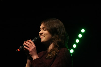 A woman sings into a microphone with a joyful expression on her face, her curly hair falling naturally over her shoulders. The background is dark, with prominent green lights forming a diagonal line behind her.