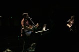 A male musician is playing a guitar and singing into a microphone on a dimly lit stage. A female vocalist stands nearby, also holding a microphone. There is a laptop on a stand in front of the guitarist.