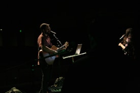 A male musician is playing a guitar and singing into a microphone on a dimly lit stage. A female vocalist stands nearby, also holding a microphone. There is a laptop on a stand in front of the guitarist.