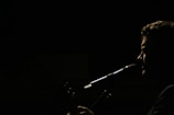 A silhouette of a person is captured in profile against a dark background, with light emphasizing their facial features and a microphone positioned near their mouth, suggesting they are singing or speaking.