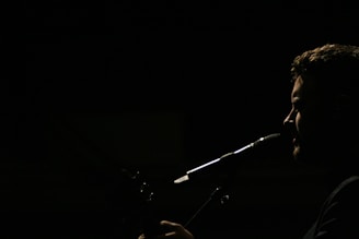 A silhouette of a person is captured in profile against a dark background, with light emphasizing their facial features and a microphone positioned near their mouth, suggesting they are singing or speaking.